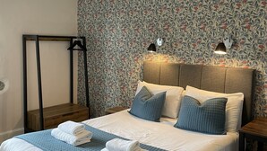 A neatly made bed with a blue quilt, white pillows, and a wooden headboard. There are two bedside tables with lamps, a hanging coat rack, and a floral-patterned wallpaper. - Wheatsheaf (Carnforth)