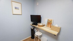 Una habitación pequeña con escritorio, televisor, cesta, taburete y cubo de basura.