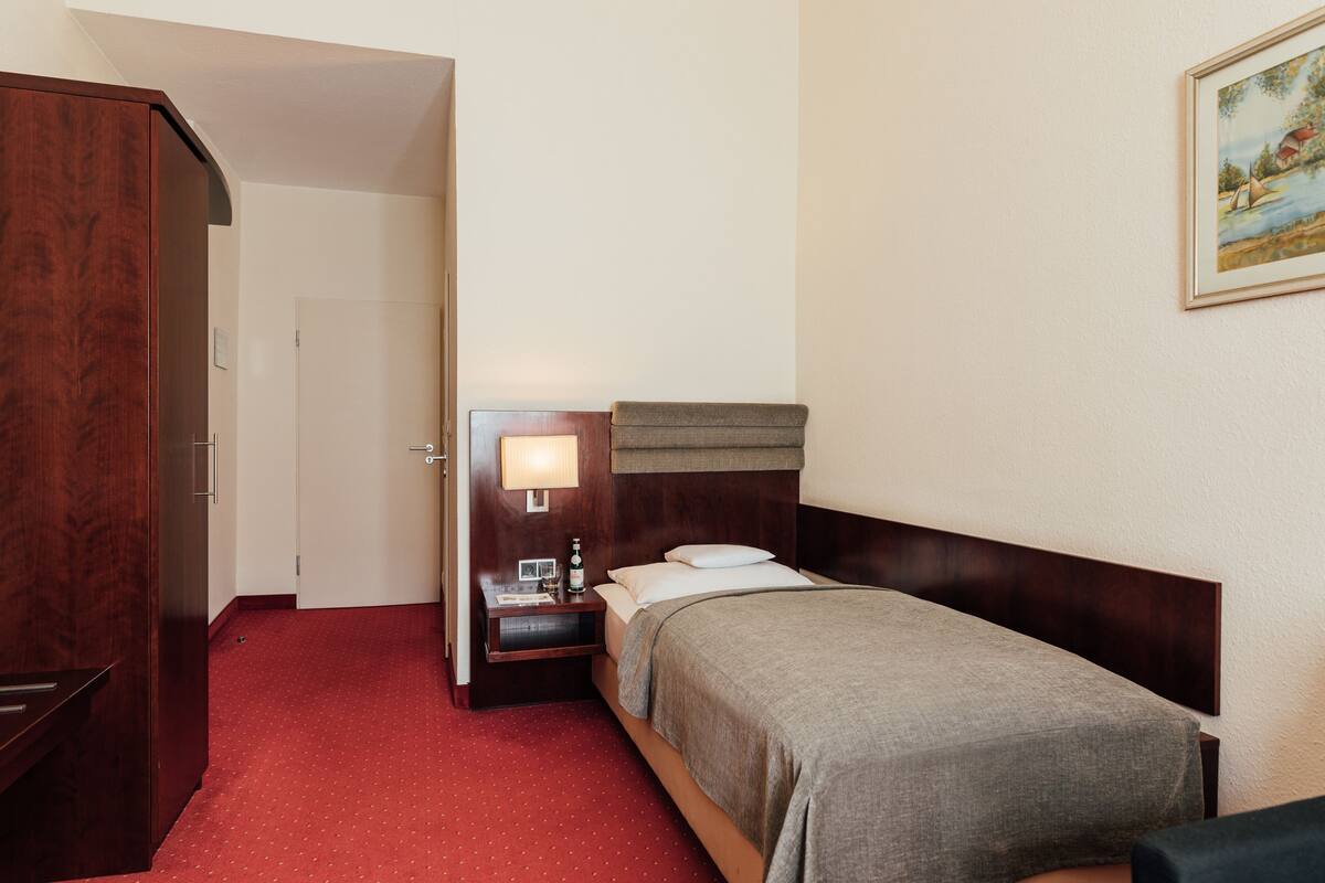 A hotel room with a single bed, a nightstand with a lamp, a wooden wardrobe, and a painting on the wall.