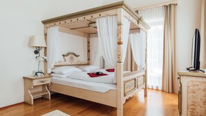 Una cama con dosel, ropa de cama blanca y estructura de madera, una mesita de noche con lámpara y un televisor sobre un mueble.