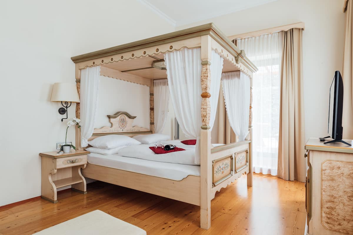 A four-poster bed with white bedding and a wooden frame, a bedside table with a lamp, and a television on a cabinet.