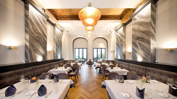 Un comedor formal con varias mesas, cada una con mantel blanco, cubiertos y copas. La habitación presenta un suelo con patrón de espiga, detalles de pared de mármol y una gran lámpara de araña.
