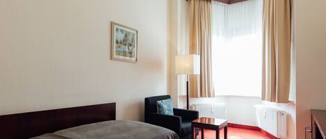 Une chambre d’hôtel avec un lit, une chaise, une petite table, une lampe, un radiateur et une fenêtre avec des rideaux.