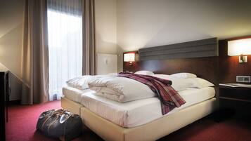 A hotel room with a bed, a nightstand, a lamp, a suitcase, and a window with curtains.