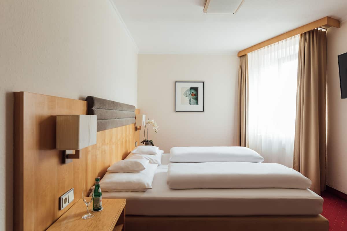 A hotel room with a bed, a bedside table with a bottle and glass, a wall-mounted lamp, a framed picture, and a window with curtains.