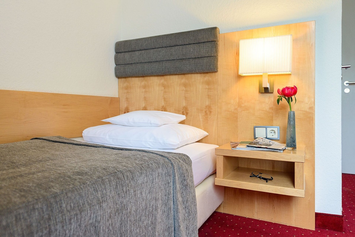 A hotel room with a bed, a bedside table with a lamp, a vase with a flower, and a small shelf with glasses.