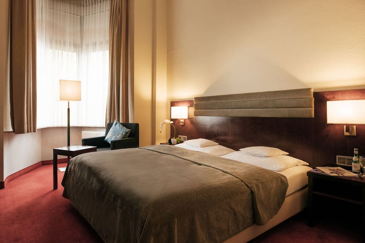 A hotel room with a large bed, a bedside table, a lamp, and a window with curtains.