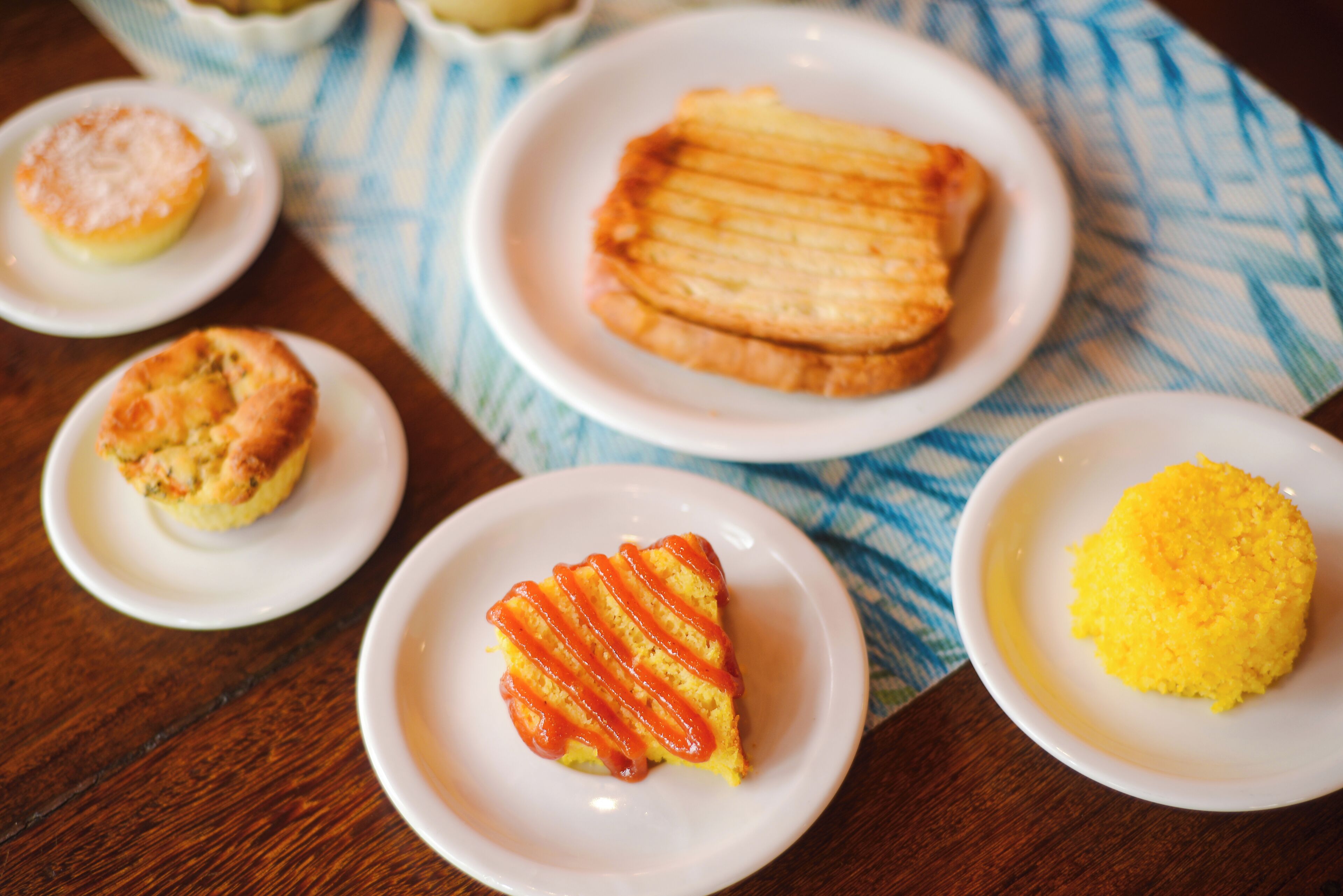 Uma mesa com diversos alimentos em pratos brancos, incluindo uma fatia de bolo com listras vermelhas, um muffin e uma sobremesa amarela.