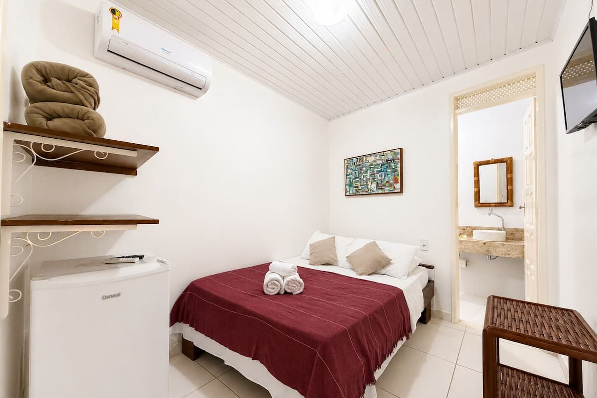 A bedroom with a bed, a bedside table, a chair, a wall-mounted air conditioner, a shelf with a towel, and a painting.