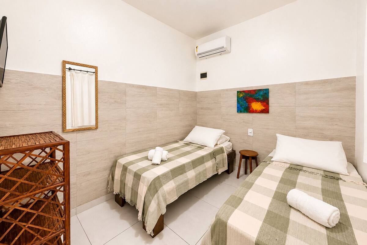 A hotel room with two beds, a small table, a chair, an air conditioning unit, and a colorful artwork on the wall.