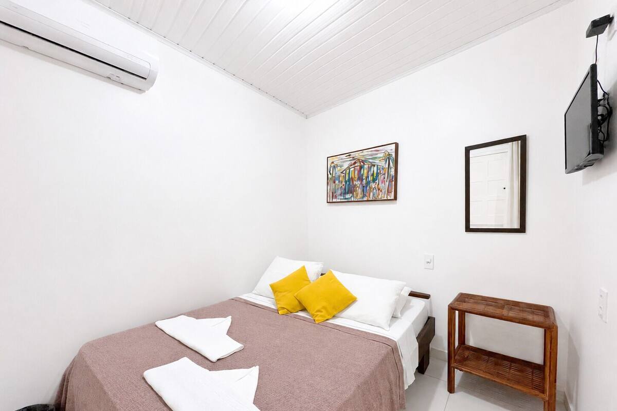 A bedroom with a bed, pillows, a bedside table, a painting, a mirror, and an air conditioning unit.