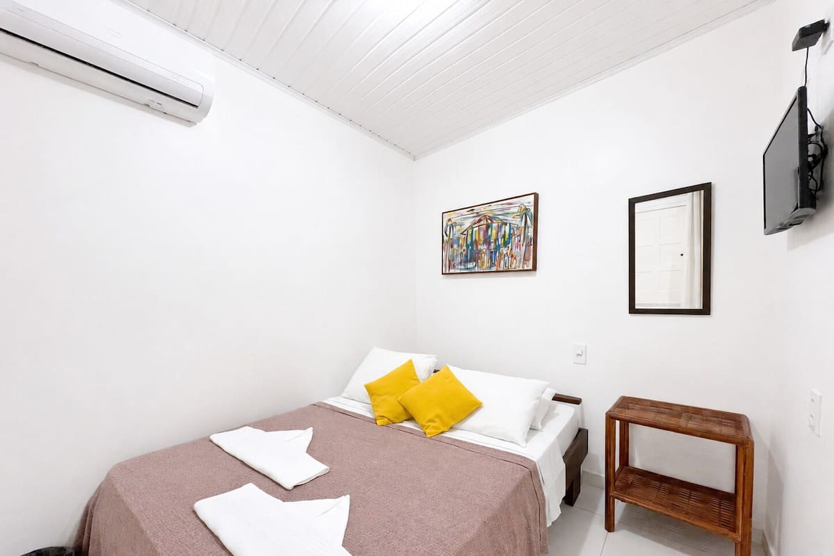 Un dormitorio con cama, almohadas, una mesita de noche, un cuadro, un espejo y un equipo de aire acondicionado.