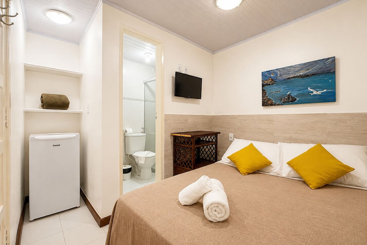 A hotel room with a bed, a television, a painting, a small refrigerator, and a shower.