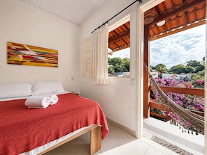 Ein Schlafzimmer mit Bett, einem Vorhangfenster, einer Hängematte und Blick auf Grünflächen und Gebäude.