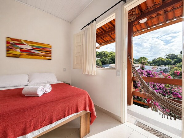 A bedroom with a bed, a window with curtains, a hammock, and a view of greenery and buildings. - Pousada Aconchego (Cairu)