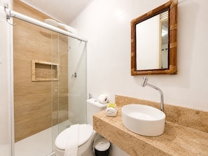 A modern bathroom with a glass shower enclosure, a toilet, a sink with a round basin, and a mirror with a wooden frame. - Pousada Aconchego (Cairu)
