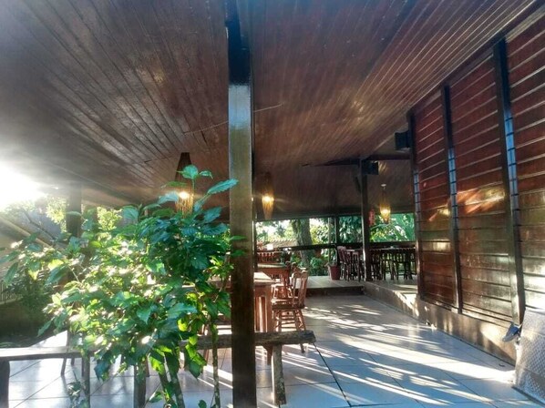 Un área de patio al aire libre con mesas y sillas de madera, rodeada de follaje y una estructura de techo de madera.