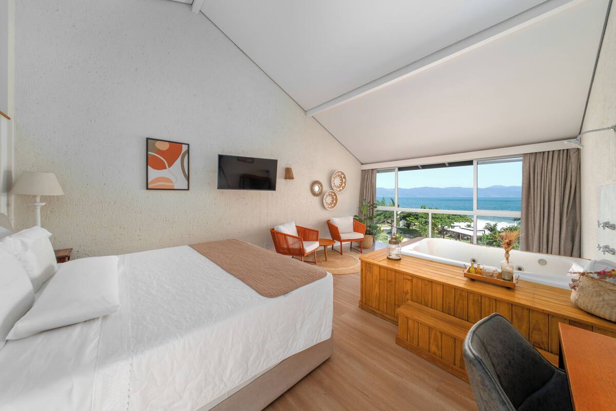 Quarto de hotel moderno com uma cama grande, uma escrivaninha de madeira, um espaço de estar com cadeiras, uma banheira e uma janela ampla com vista para o mar.
