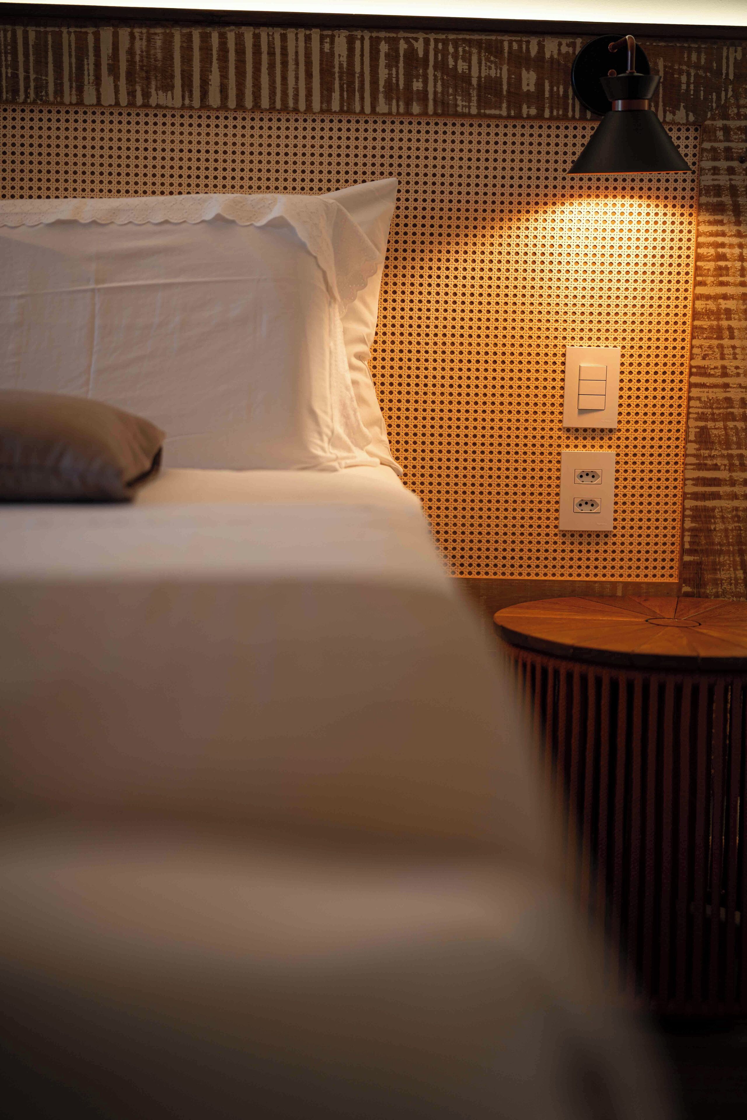Una cama bien tendida con sábanas blancas y un almohadón, una mesita de noche redonda de madera con una lámpara, y una pared con un diseño perforado.