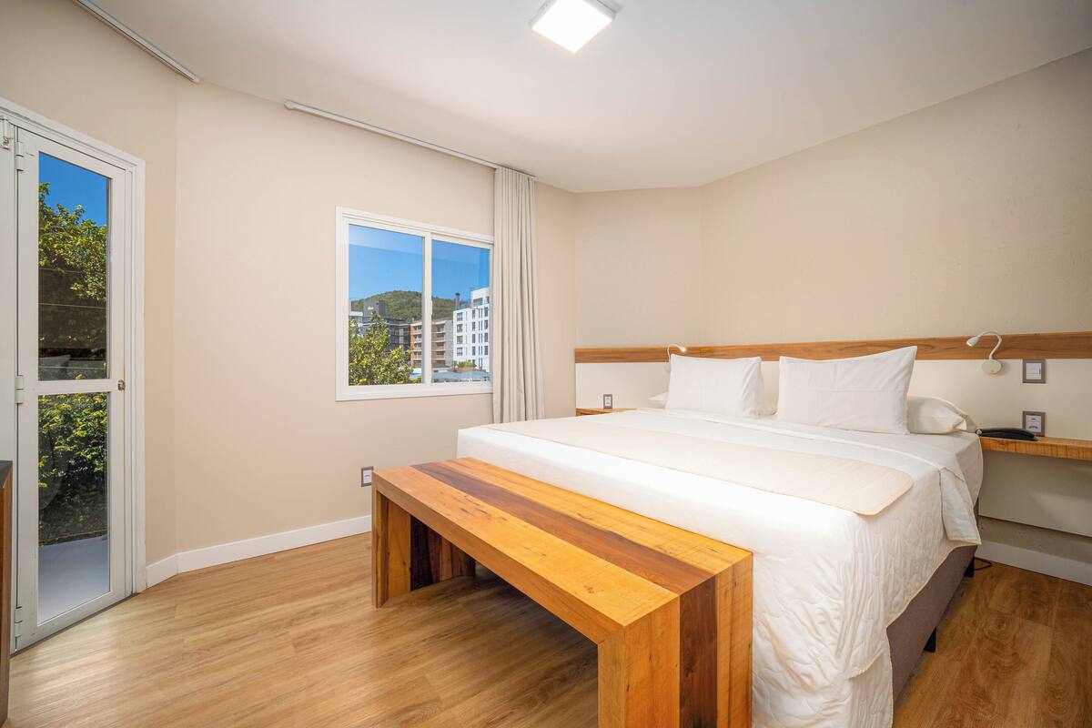 Quarto de hotel moderno com uma cama grande, um banco de madeira, uma janela com vista para a cidade e uma porta que dá para uma área externa.