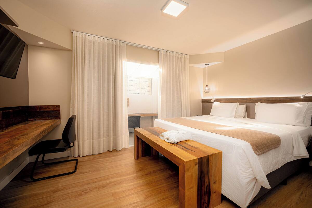 Quarto de hotel com escrivaninha de madeira, cama com roupa de cama branca, um banco de madeira e uma janela grande com cortinas.