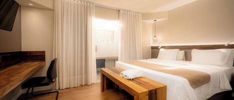 A hotel room with a wooden desk, a bed with white bedding, a wooden bench, and a large window with curtains.