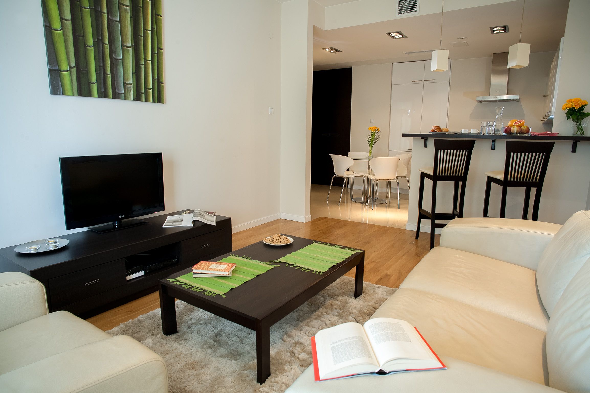 Una sala moderna con un televisor de pantalla plana, una mesa de centro de madera oscura, un sofá blanco y un comedor con dos sillas y una mesita.