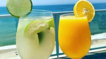 To cocktails med lime og appelsinpynt, holdt i hænder, med udsigt til havet i baggrunden.