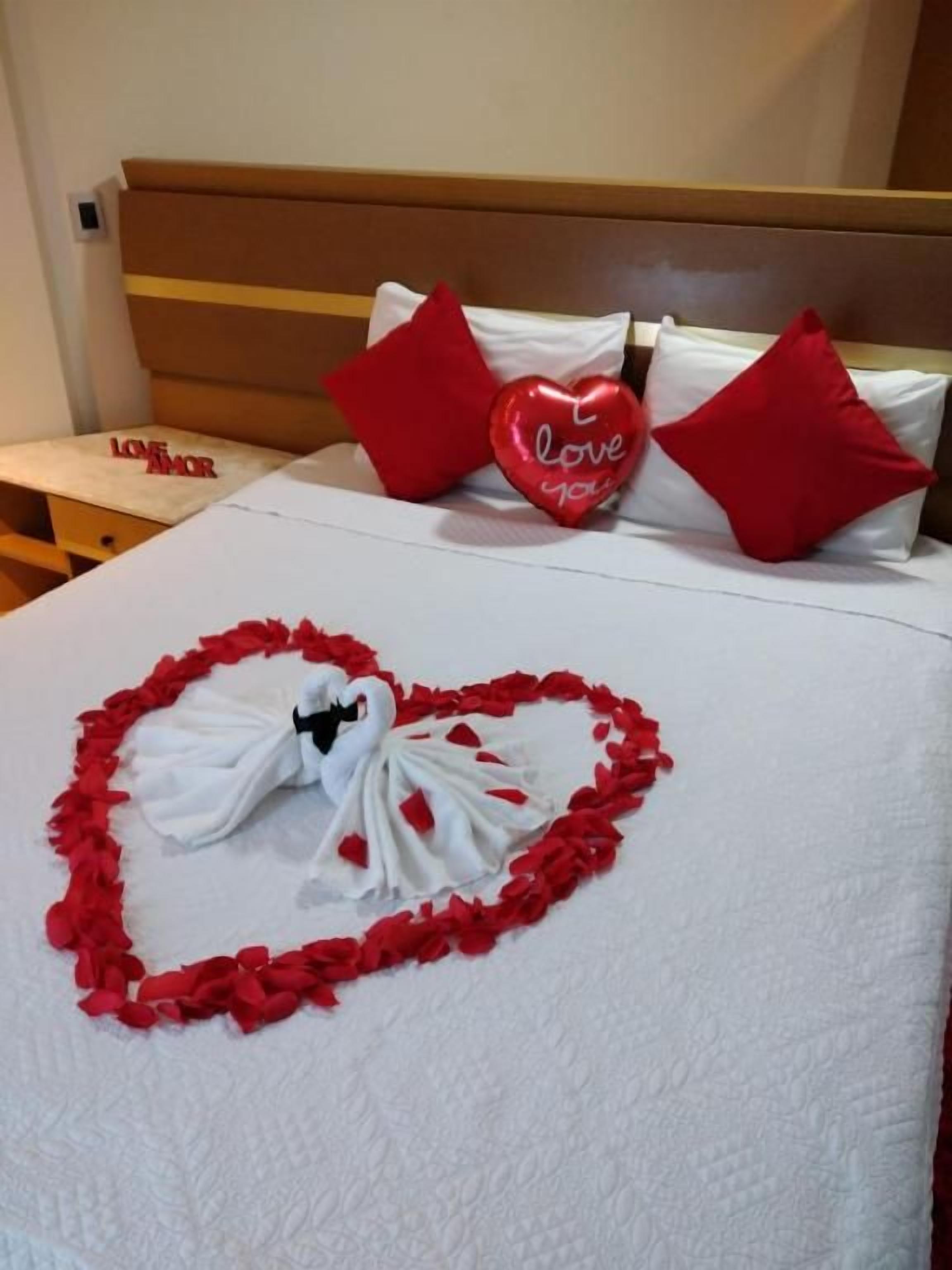 Una cama con una toalla dispuesta en forma de corazón y almohadas con la inscripción 'love you'.