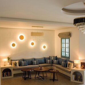 A cozy living room with a sectional sofa, round tables, and a large window with curtains. - Armonia Hotel (Santorini)