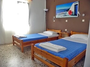 A room with two wooden beds, each with a blue bedspread and white pillows. There is a painting of a coastal scene on the wall, a floor lamp, and a window with curtains. - Armonia Hotel (Santorini)