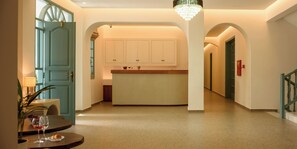 A spacious hallway with a dining area featuring a table with a glass of red wine and pastries. There is a kitchen area with white cabinets and a chandelier. - Armonia Hotel (Santorini)
