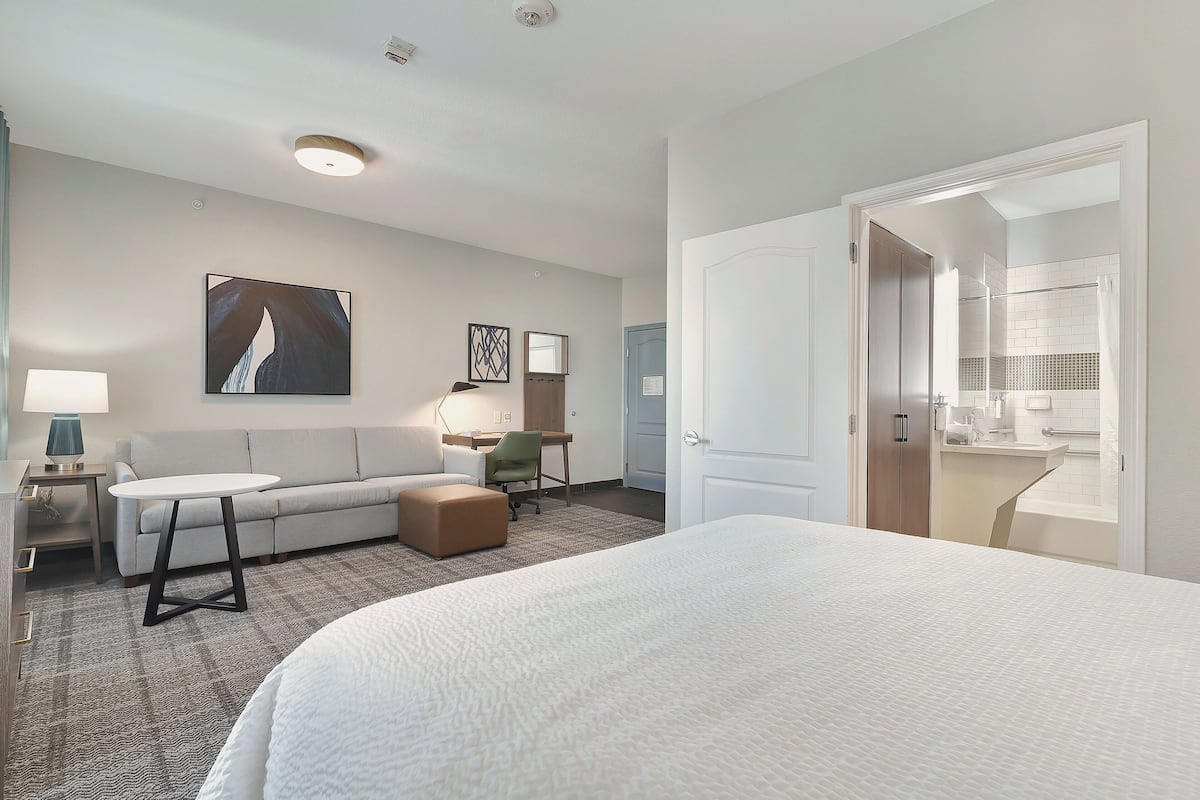 A hotel room with a large bed, a sofa, a desk, and a bathroom visible through an open door.