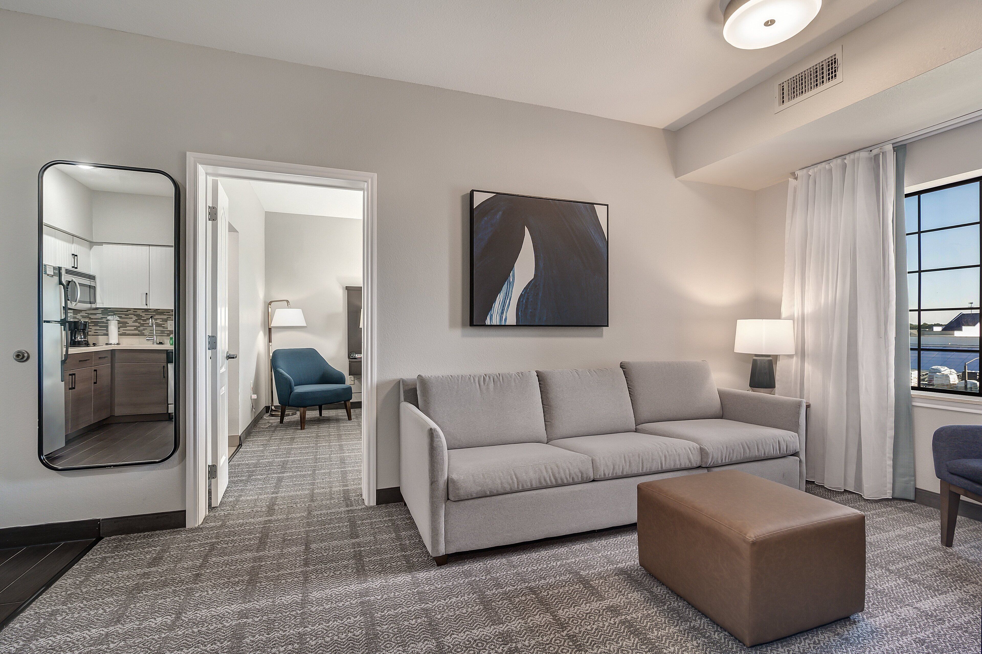 A hotel room with a grey sofa, a brown ottoman, a blue armchair, a large mirror, and a painting on the wall.