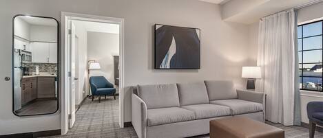 A hotel room with a grey sofa, a brown ottoman, a blue armchair, a large mirror, and a painting on the wall.