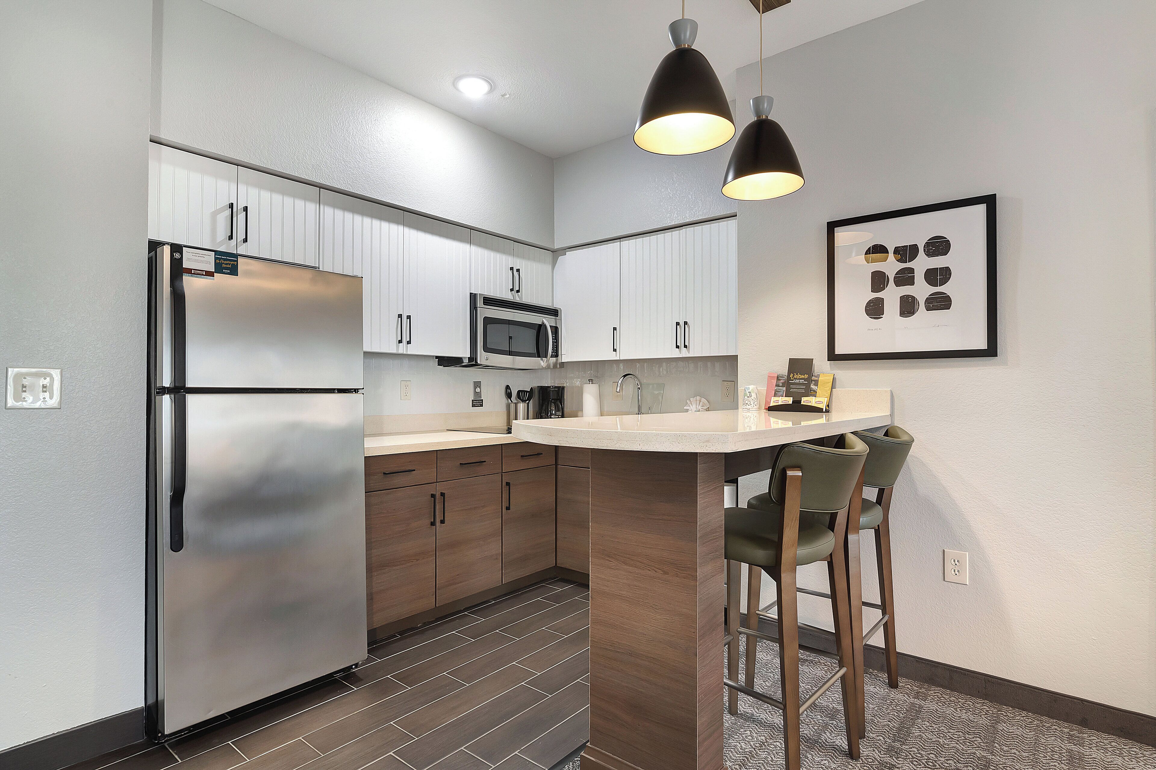 A modern kitchen with a stainless steel refrigerator, microwave, and wooden cabinets. There is a bar area with two stools.