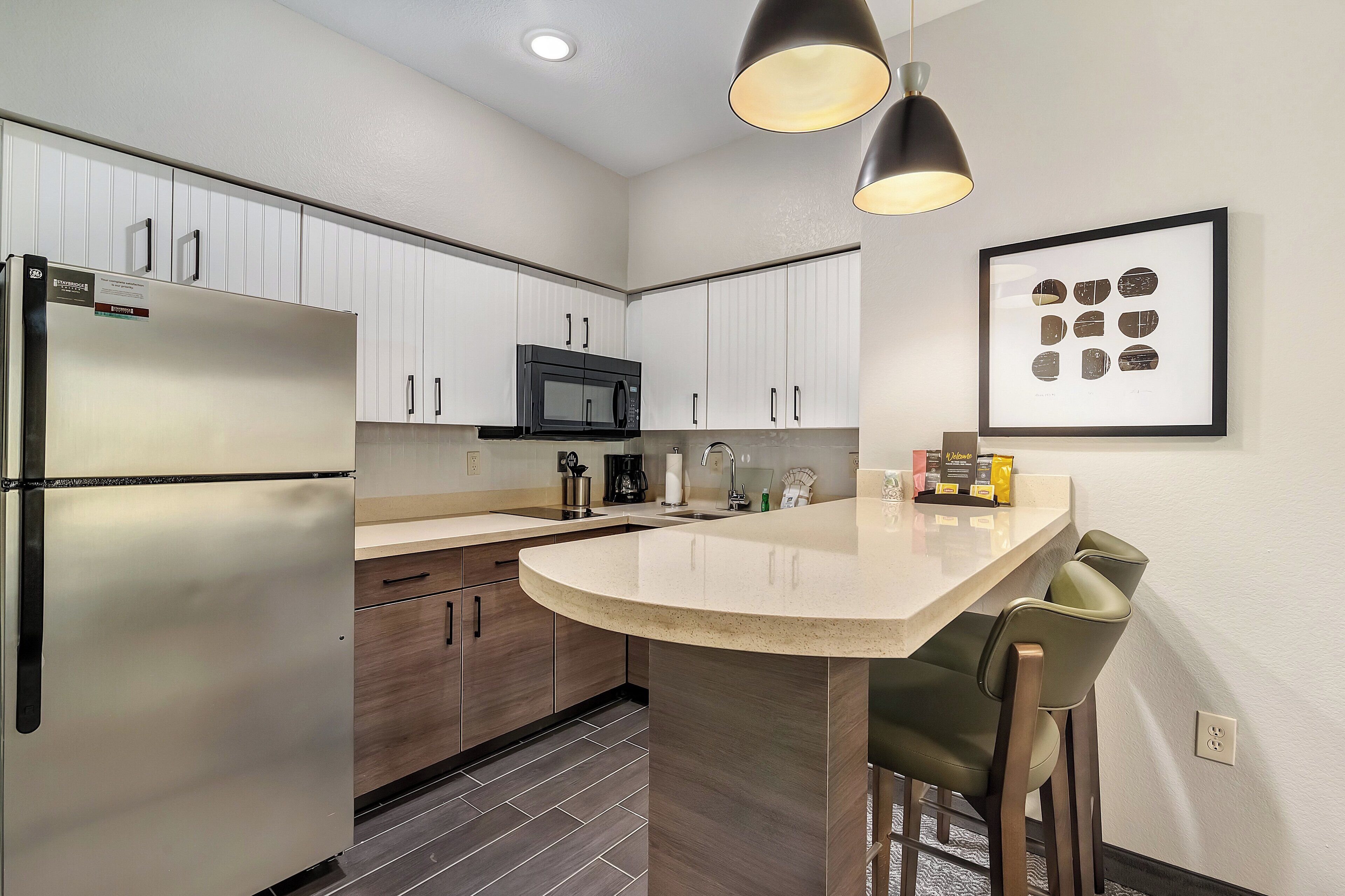 A modern kitchen with a stainless steel refrigerator, a microwave, and a sink. There is a framed picture on the wall and a bar area with two chairs.