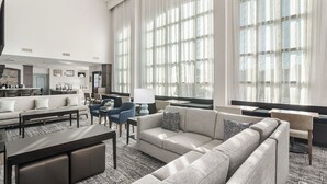 Un amplio vestíbulo de hotel con una zona de estar grande, una cocina al fondo y ventanas grandes que dejan entrar luz natural.