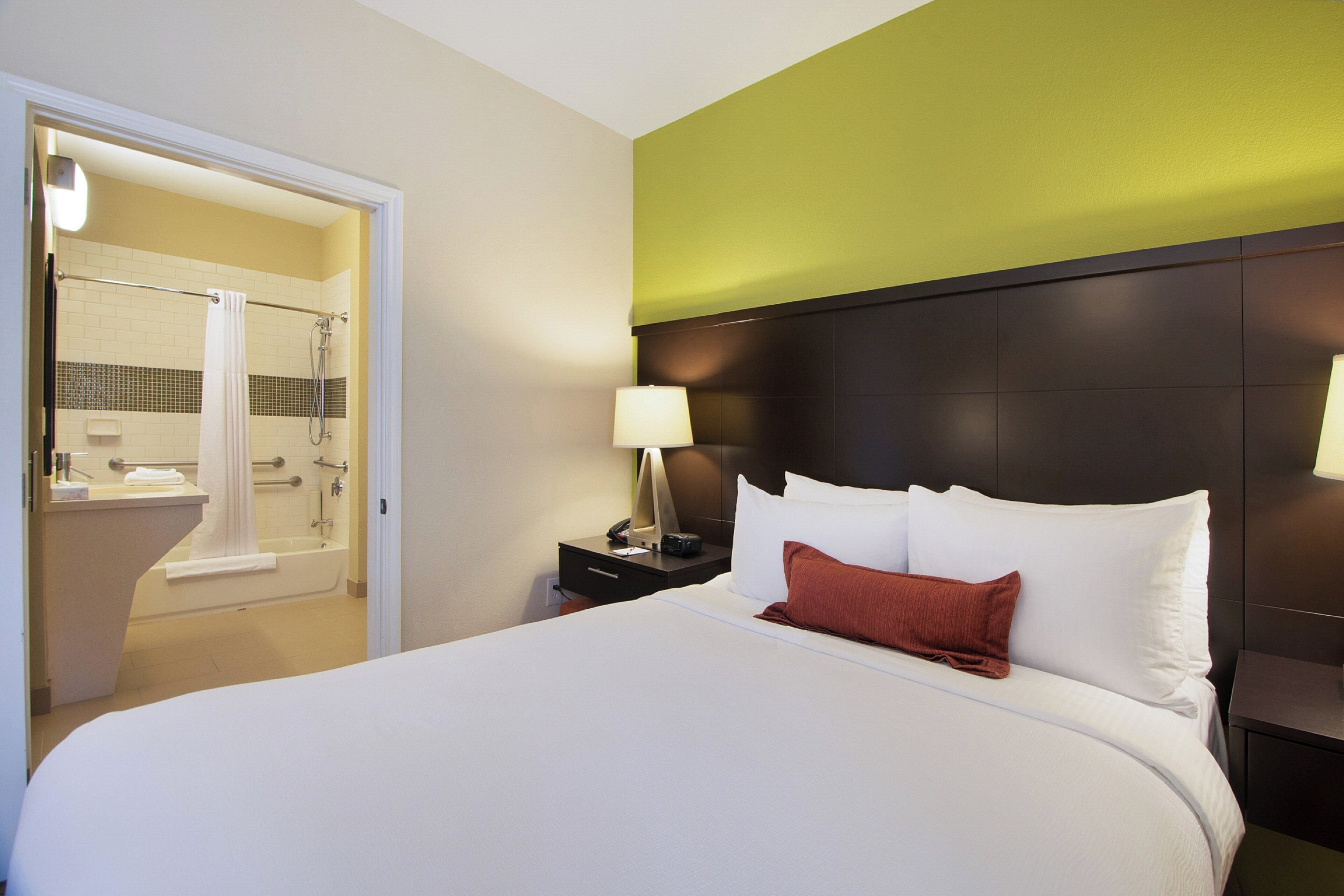 A hotel room with a bed, a nightstand, a lamp, a bathroom with a shower, and a door leading to another room.