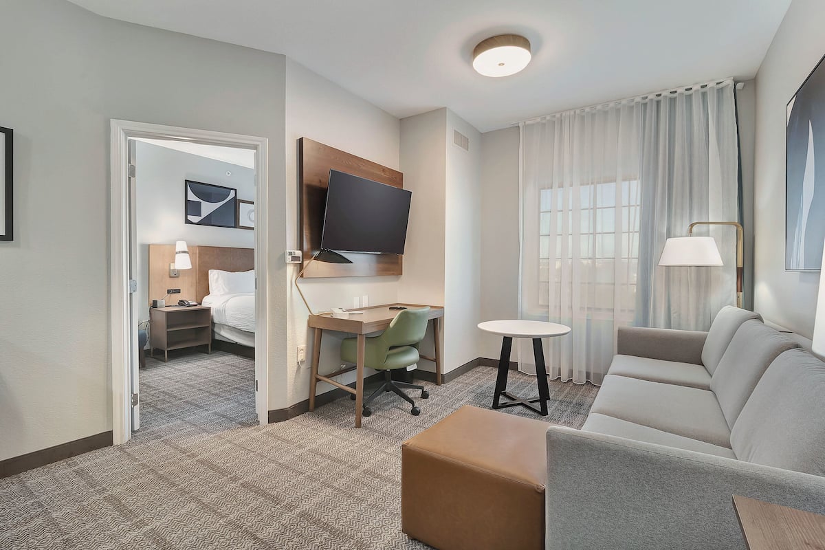 A hotel room with a sofa, a TV mounted on the wall, a desk with a chair, a small round table, and a bed in the adjacent room.