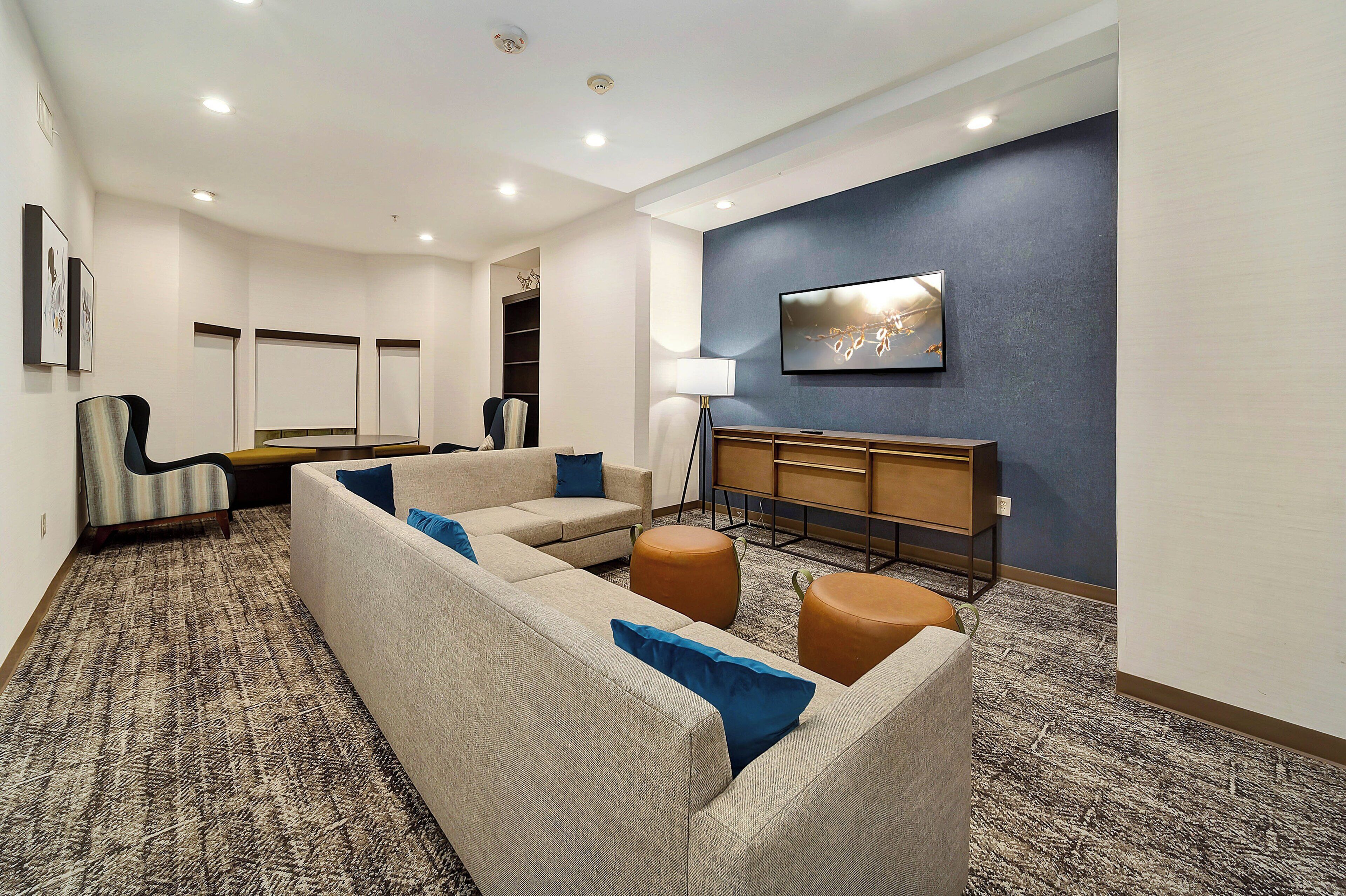 A hotel room with a large sectional sofa, a small round ottoman, a desk with a lamp, and a flat-screen TV mounted on a dark wall.