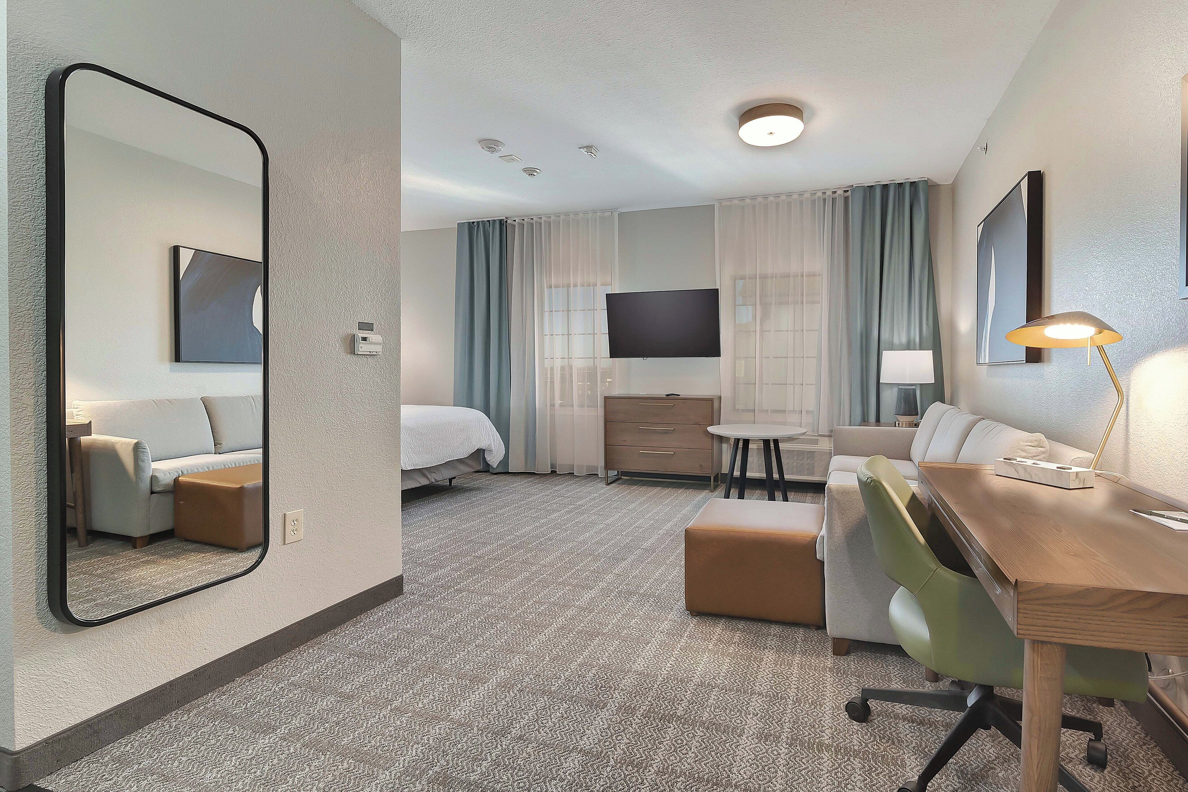 A hotel room with a bed, a desk, a chair, a TV, a lamp, and a large mirror.