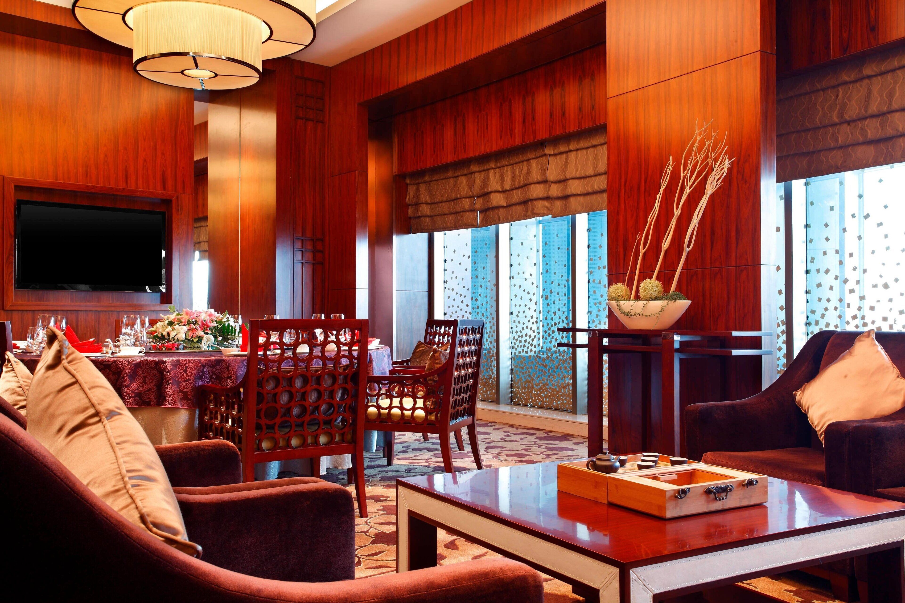 A hotel room with a wooden interior, a flat-screen TV mounted on the wall, a dining area with a table set for two, and a seating area with a sofa and armchairs.