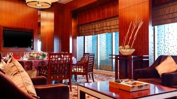 A hotel room with a wooden interior, a flat-screen TV mounted on the wall, a dining area with a table set for two, and a seating area with a sofa and armchairs.