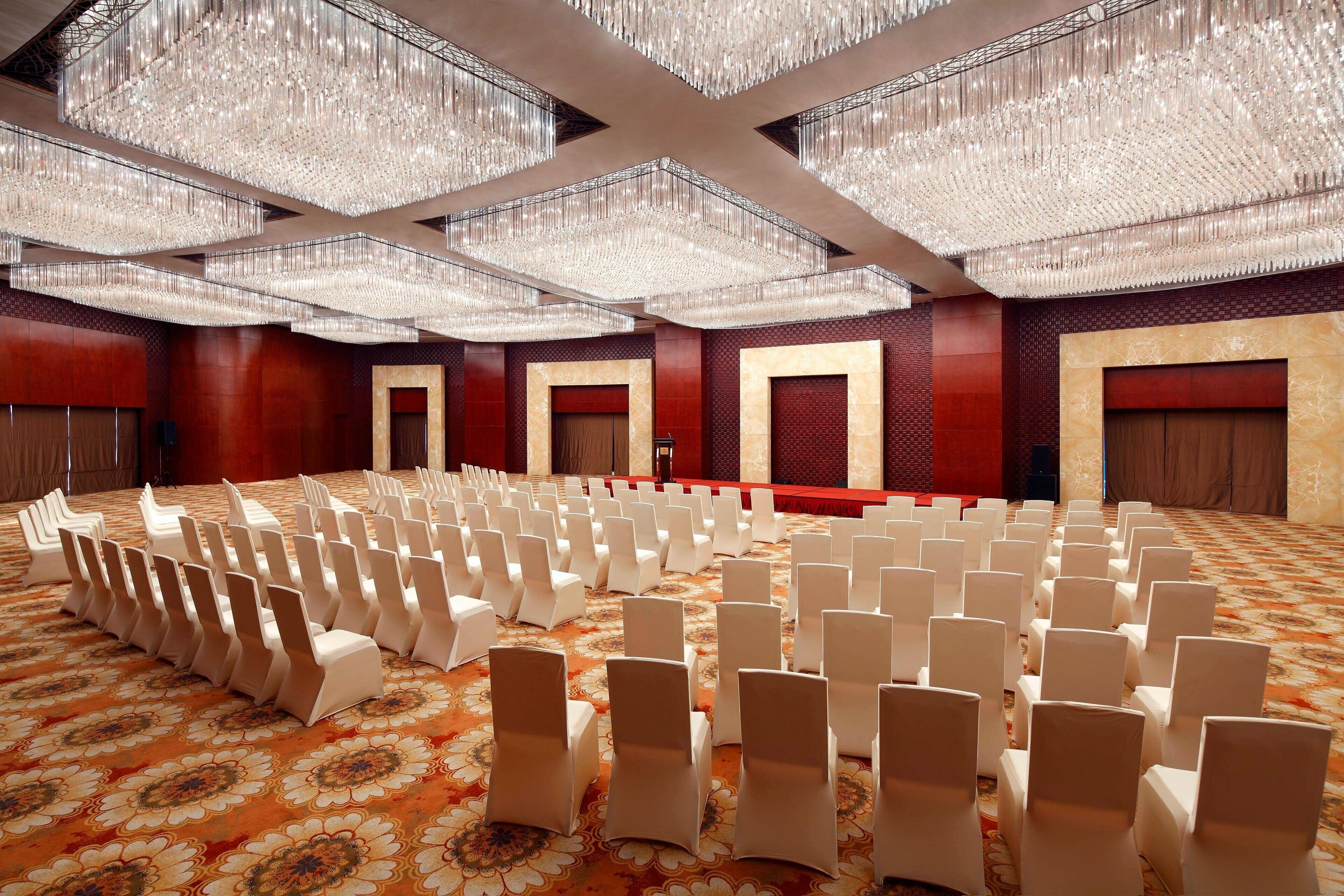A large hall with rows of white chairs, a patterned carpet, and a ceiling with multiple chandeliers.
