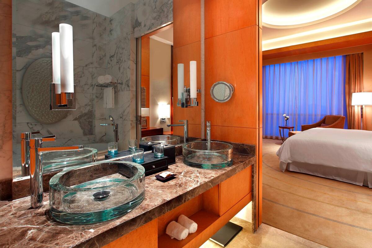 Une salle de bain moderne avec un grand miroir, deux vases en verre posés sur le lavabo, et une vue sur une chambre à coucher avec un lit et un rideau bleu.
