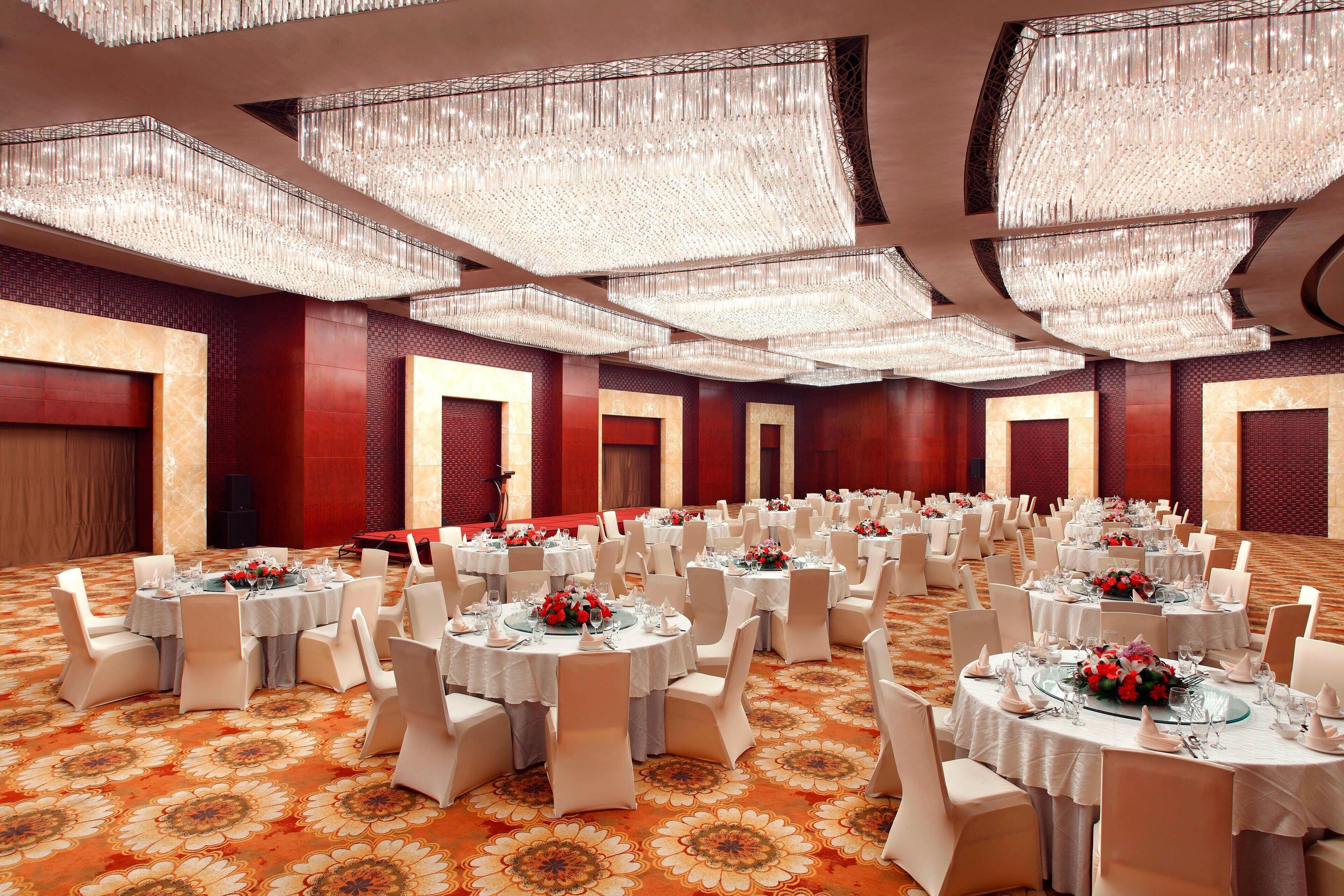 A banquet hall with round tables set for events, featuring a patterned carpet and chandeliers.
