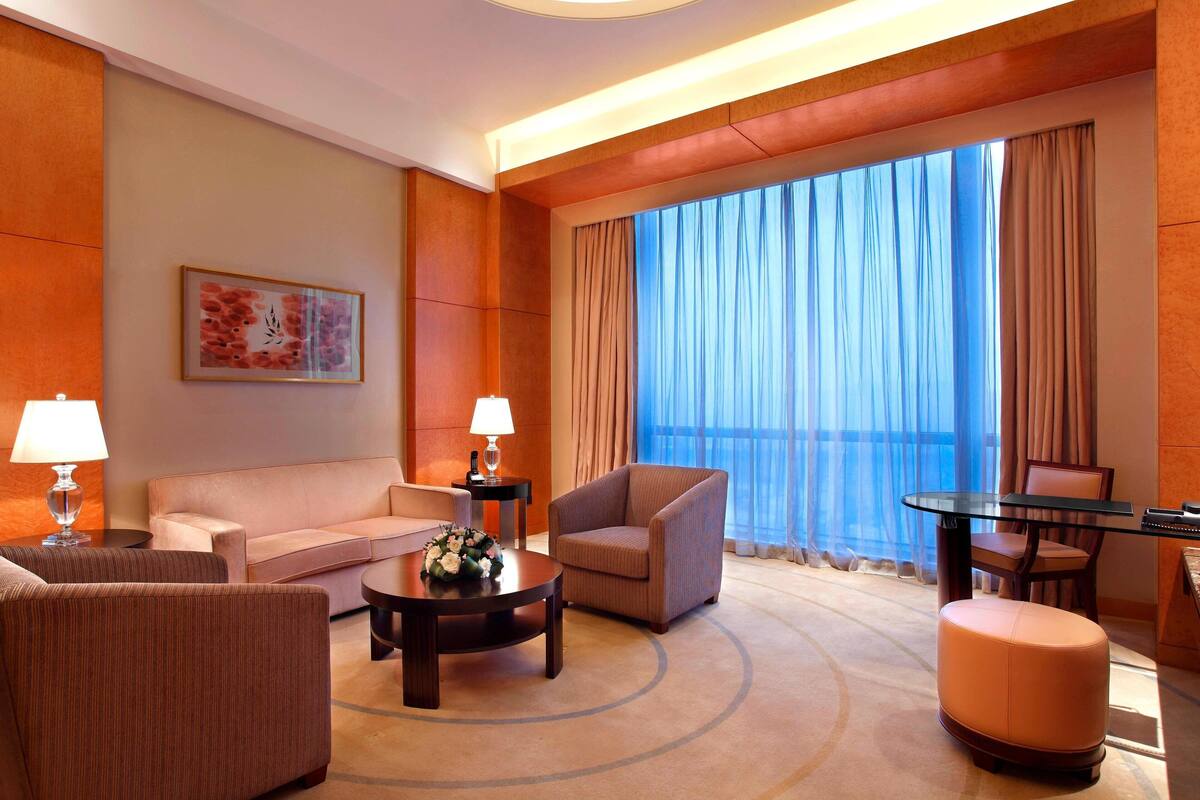Une chambre d’hôtel comprenant un canapé, un fauteuil, un pouf, une table basse et une table à manger avec des chaises. Il y a une grande fenêtre avec des rideaux, une lampe et un tableau encadré au mur.