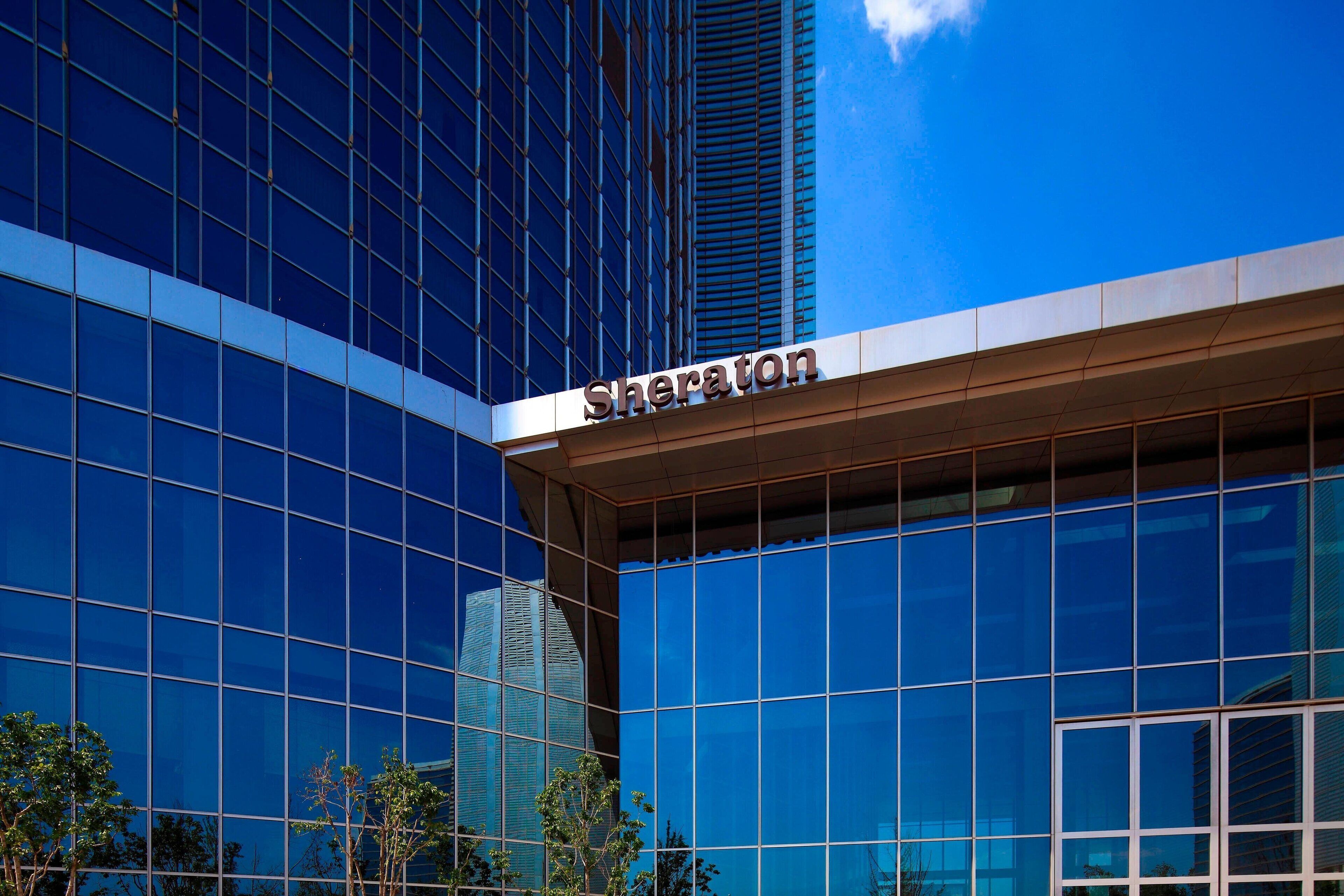 Un hôtel Sheraton doté d’une façade moderne en verre et dont les fenêtres reflètent un ciel d’un bleu limpide.