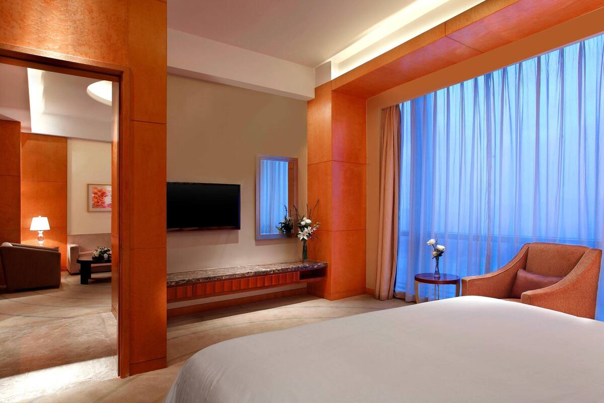Une chambre d’hôtel avec un grand lit, un téléviseur fixé au mur, une petite table d’appoint sur laquelle est posé un vase de fleurs, et une vue sur l’extérieur à travers des rideaux légers.